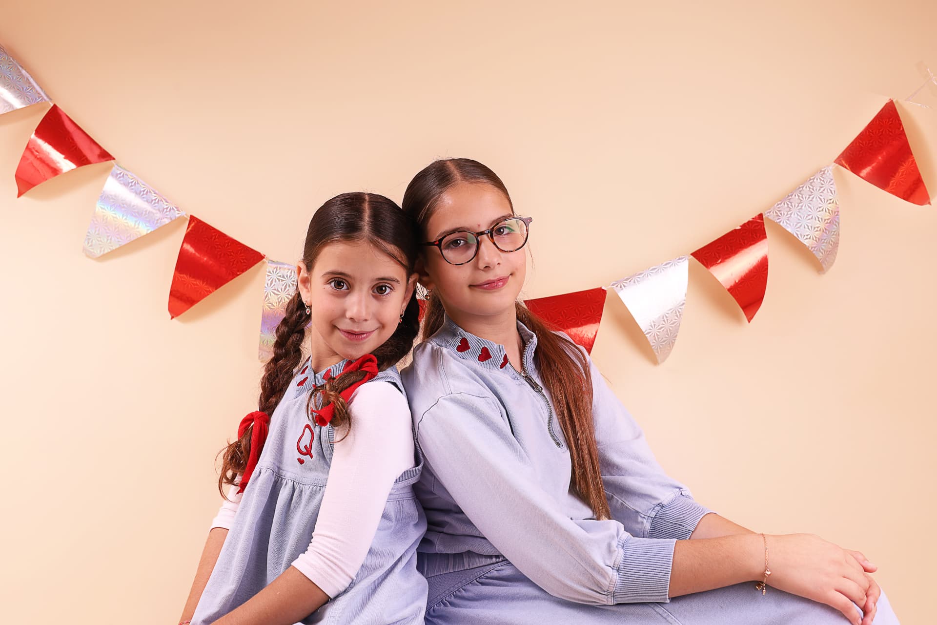 שתי אחיות יושבות גב אל גב ומחייכות מתחת לגירלנד אדום