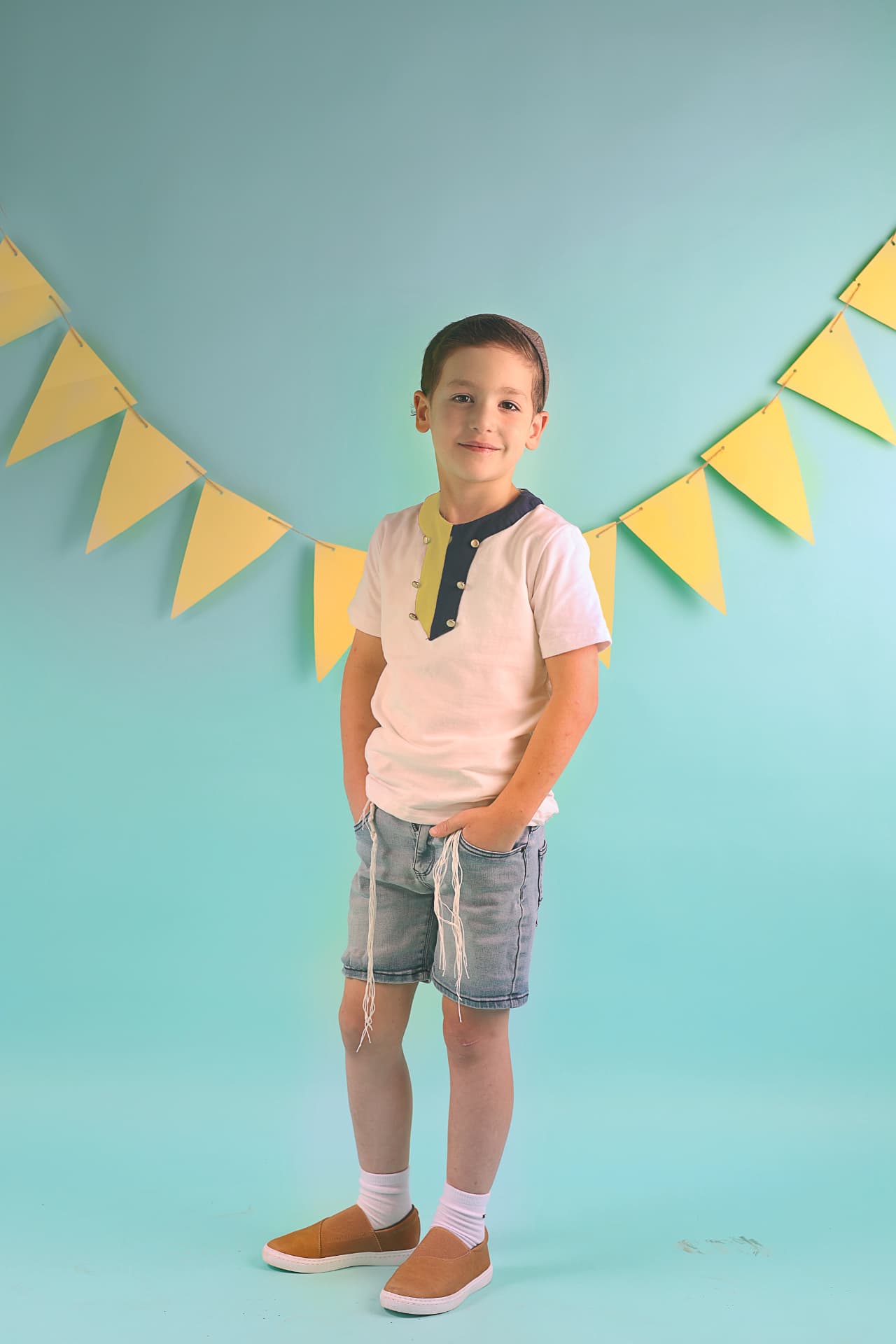 ילד עם כיפה וגרביים עומד מול רקע טורקיז עם גירלנד צהוב